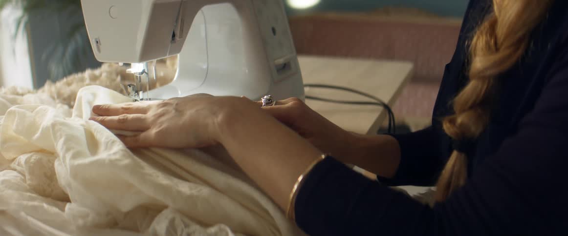 Woman with long braided hair pushes filmy fabric under sewing machine needle