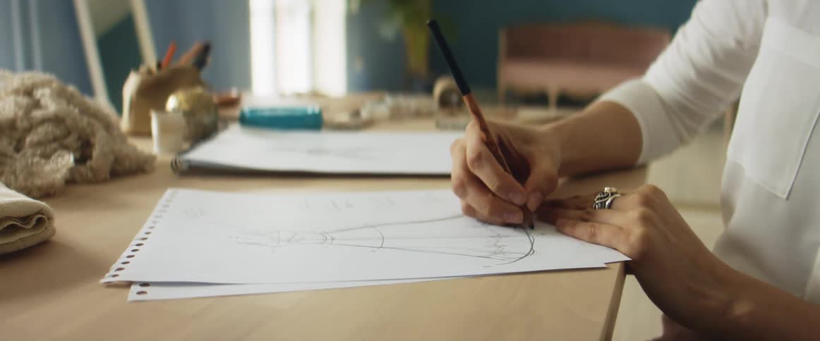 Female clothing designer sits at wooden table drawing dress