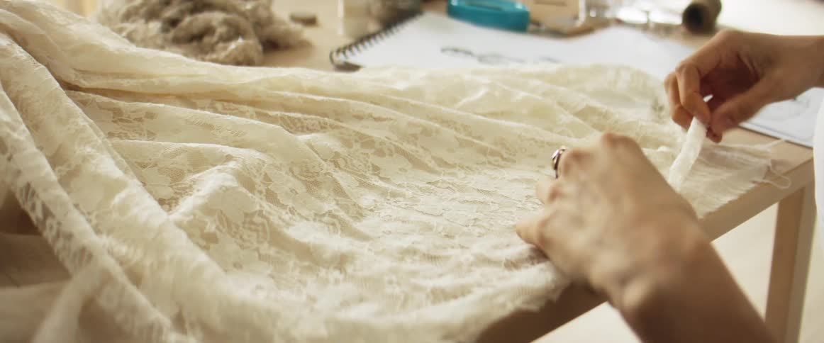 A woman smooths out lace fabric while sitting with a sketch of a dress
