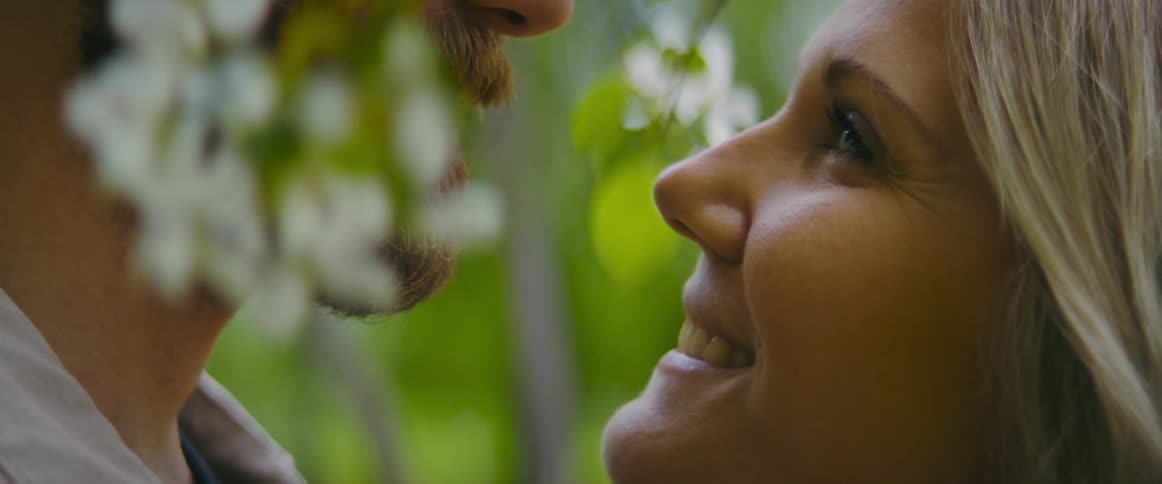 A smiling woman gazes into the eyes of a man as they embrace under a blossoming tree during the day