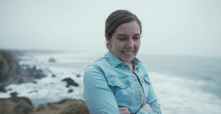 A woman standing on the shore smiles as waves crash below her