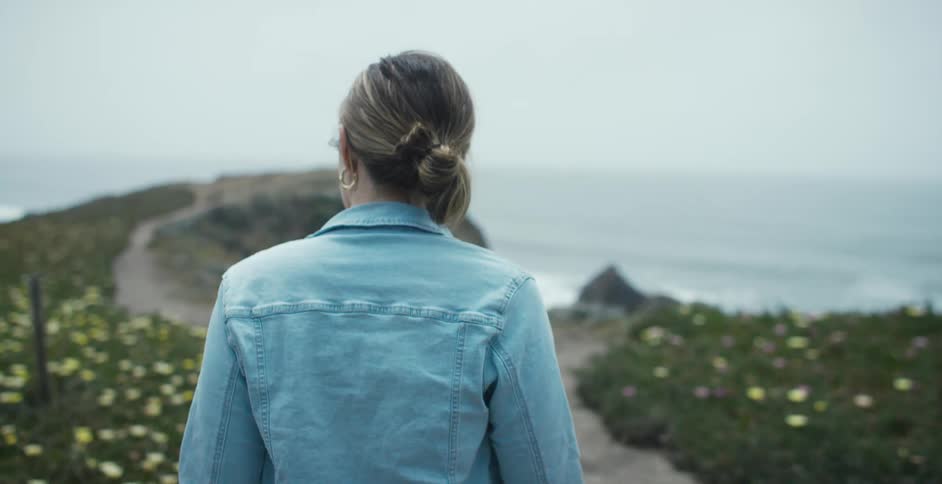 Woman wearing blue denim jacket walks on a dirt road towards the ocean