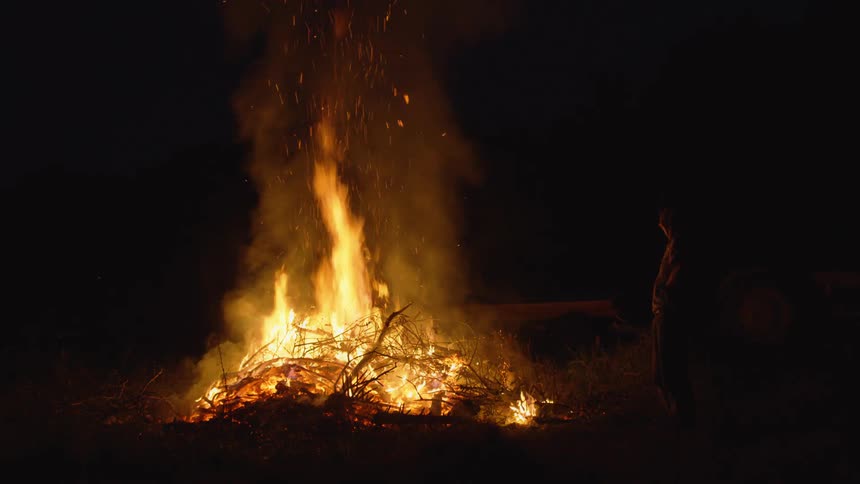 A man wearing cap approaches burning bonfire
