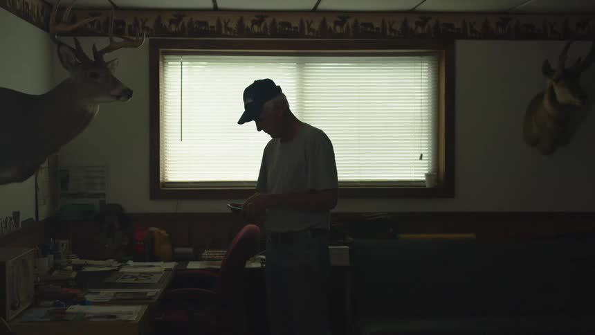 Elderly man enters a dimly lit room with hunting trophies on the walls checking his smartphone