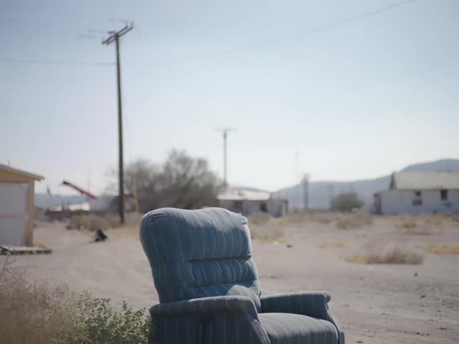 A fabric armchair sits on a dirt road next to a bush with dilapidated homes behind it