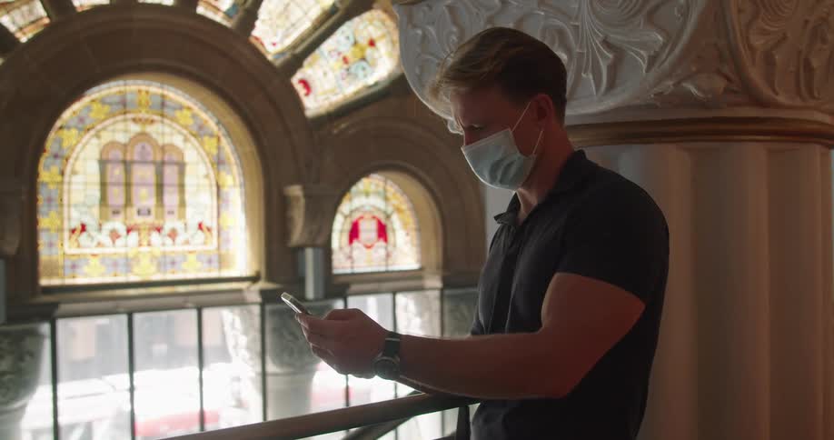 Man wearing face mask stand near column in ornately decorated room that includes elaborate stained glass windows making call on mobile phone