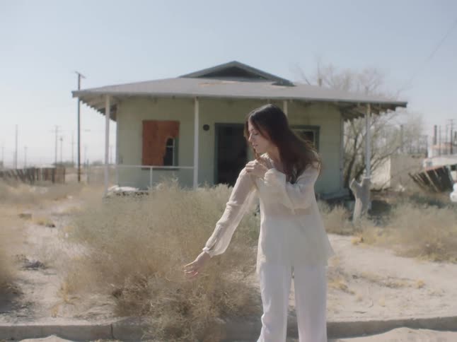 A woman dances on the street in front of a run down isolated house