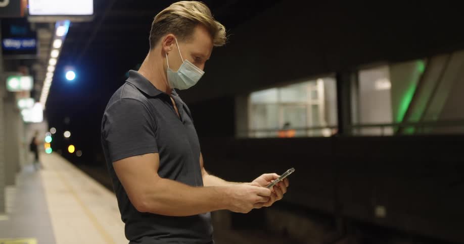 Man wearing a face mask text on his smartphone on a train station at night