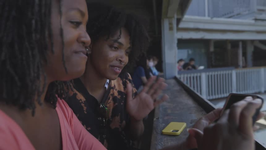 Two women on a balcony looking at a smart phone with trees the sky and clouds in the background