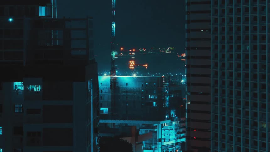 Lights shining from office at night match lights on building construction site as construction crane moves