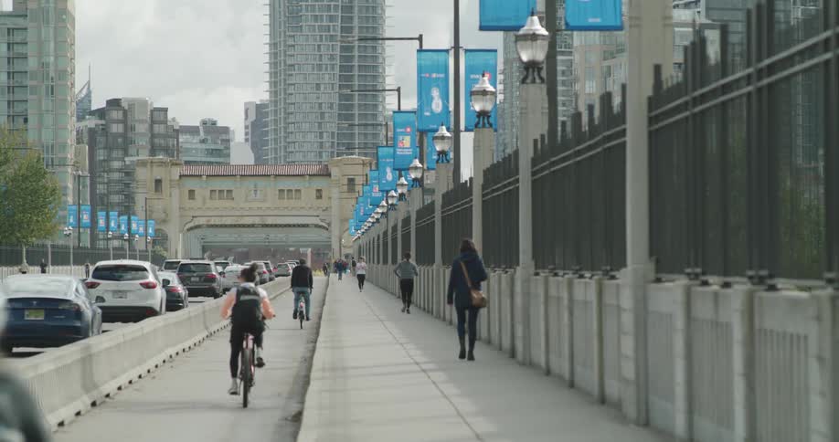 Multiple people ride a bike and walk on a large sidewalk beside a city road full of vehicles