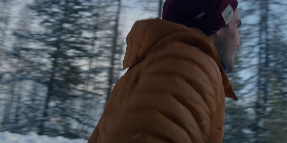 A man with a hat and coat walking on snow with trees the sky and clouds in the background