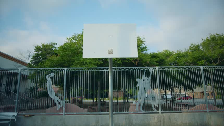 Basketball board without hoop stands next to fence containing art forms of basketball players in park
