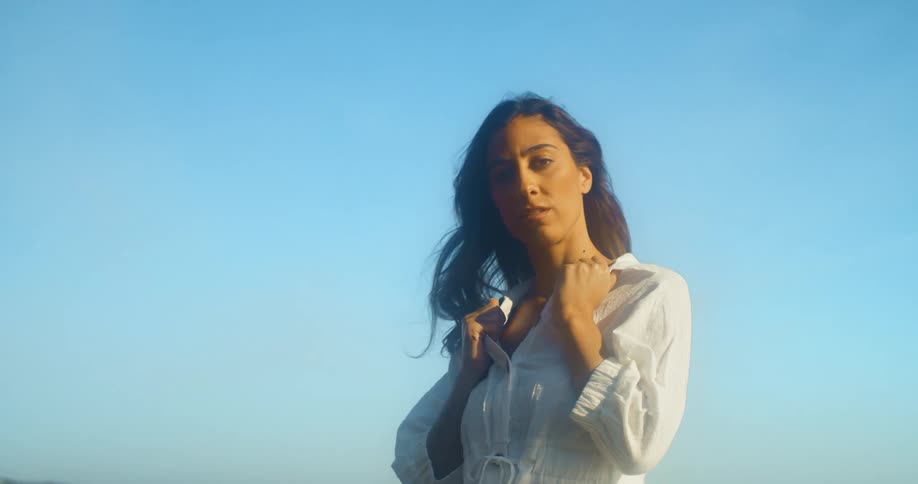 A woman stands outside and hold on to her collar and looks off in to the distance