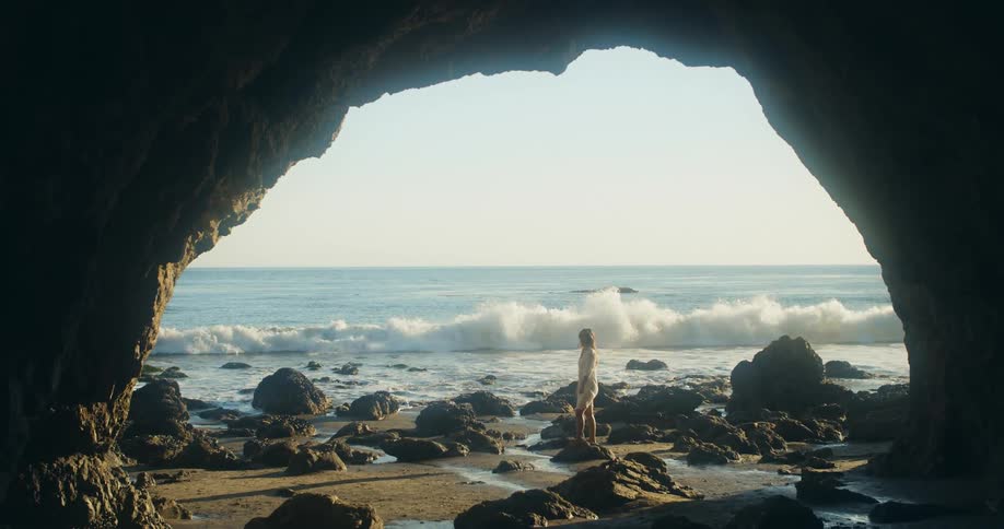 A woman stands on the rocky beach next to the ocean and a large cave