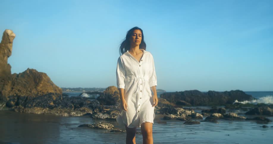 Waves splash against the rocks as a woman in a white dress walks on shore