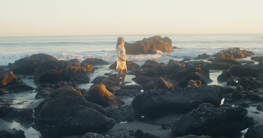 A woman standing on a rocky shore with the ocean the sky and clouds in the background