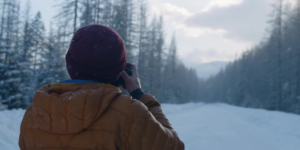 A man stands on a snow covered road lined with forest trees and takes a photo