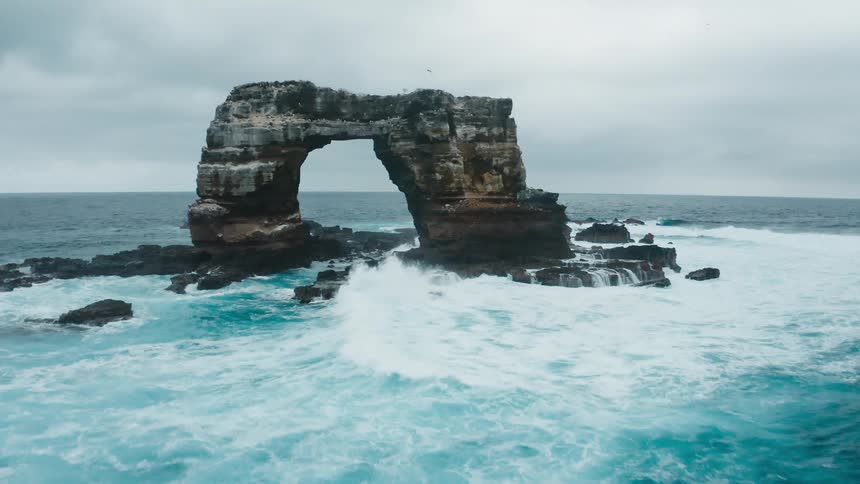 Waves splashing against a natural arch rock formation under a cloudy sky