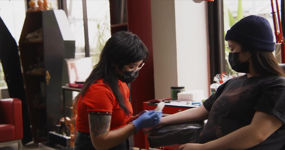 A tattoo artists uses a razor and a cloth to prepare a customers arm for a tattoo