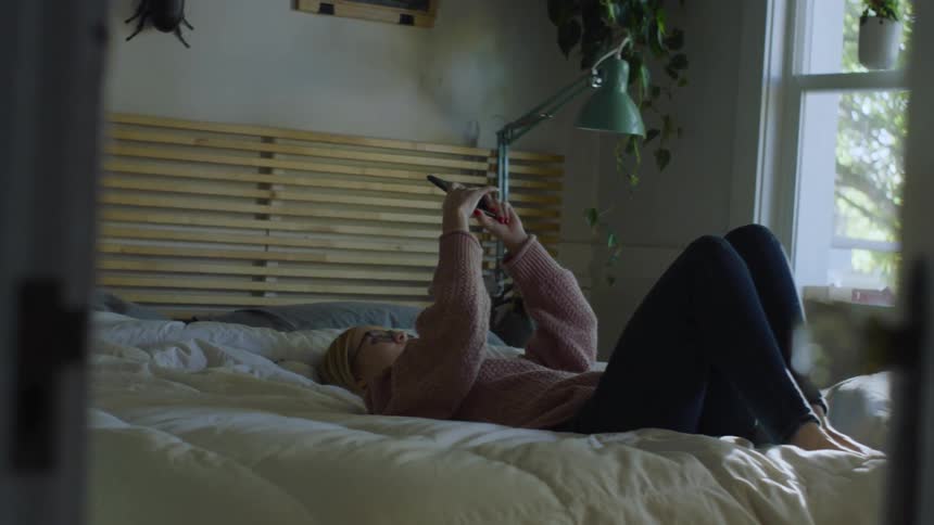 Blonde woman with pink sweater and jeans lies on bed and uses smartphone in bedroom