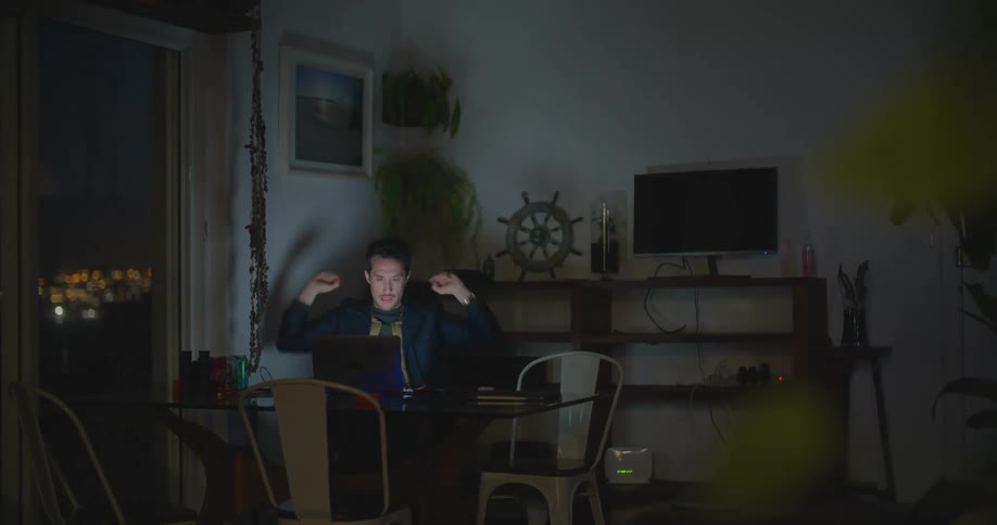 A man sits at a table in the dark with a laptop and stretches in exhaustion