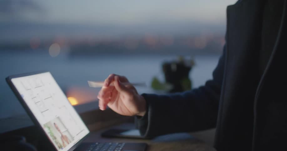 A man sits in front of a window showing a body of water and coastline while he works on a computer and laughs