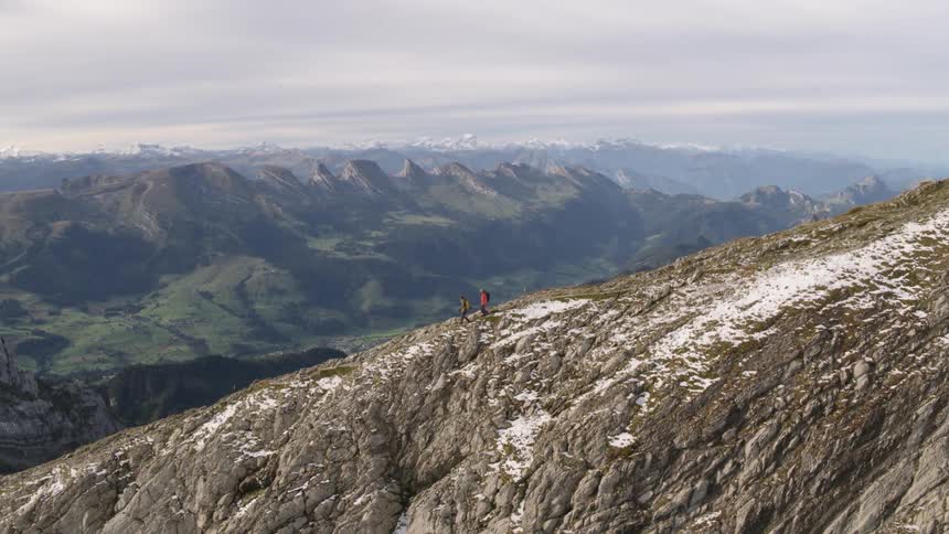 Two people walking on top of a mountain ridge with mountains in the distance on a cloudy day
