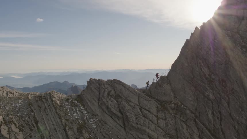 Two people climbing on top of a mountain ridge with the sun shining brightly in the sky