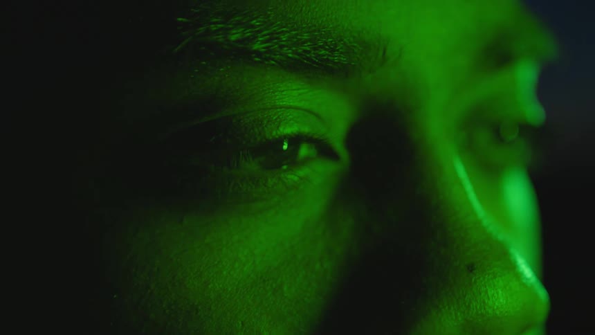 A male stares with a dull expression as a green light illuminates his face