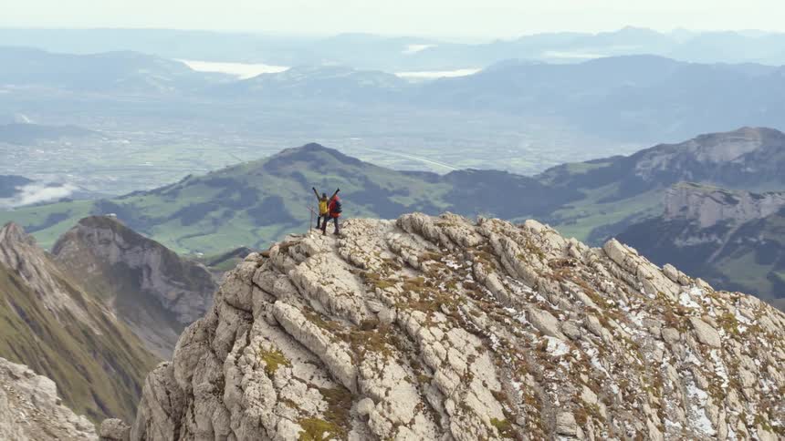 Two people standing on top of a mountain ridge with mountains in the distance on a cloudy day