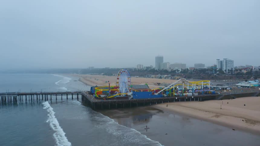 A boardwalk on a beach with an amusement park with buildings the sky and clouds in the background