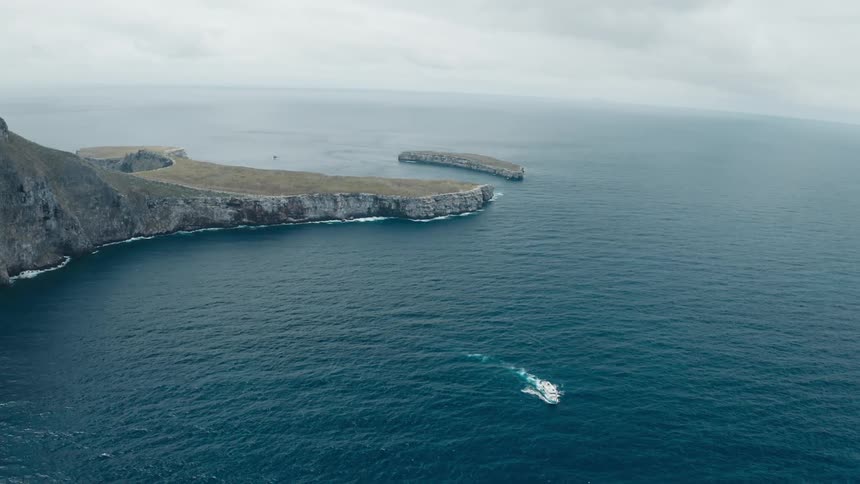 Boat cuts through water away from sheer stone of shore and promontory on overcast day