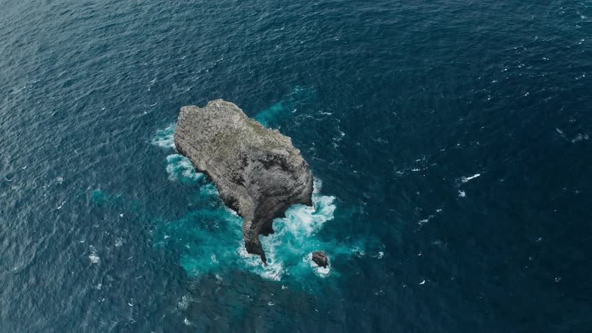 A rock formation protrudes above the ocean surface a waves break against it
