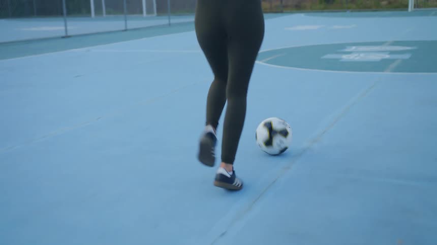 A woman in a playground kicking a soccer ball with trees the sky and clouds in the background