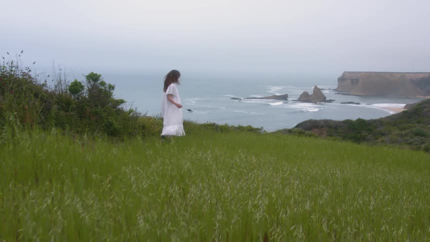 Woman with white dress walks downhill with cliffs and coast in the background