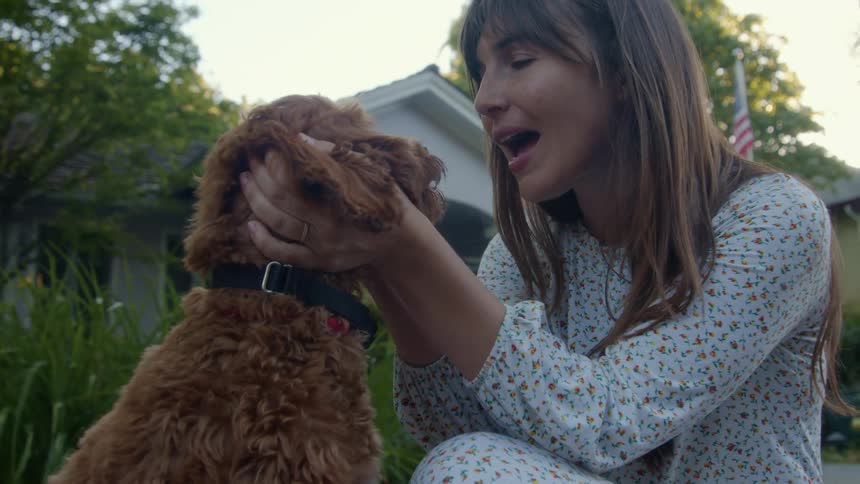 A woman sits outside a house and pets and talks to a dog