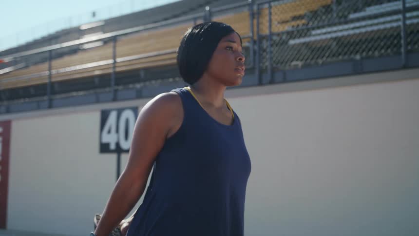 Woman stands on running track next to stadium seating stretching her leg