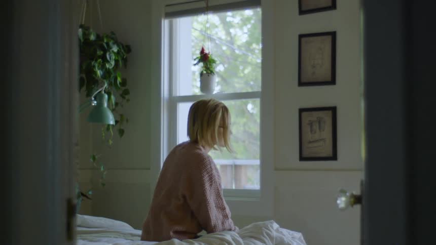 Woman brushes her hair back while she sits on the edge of an unmade bed