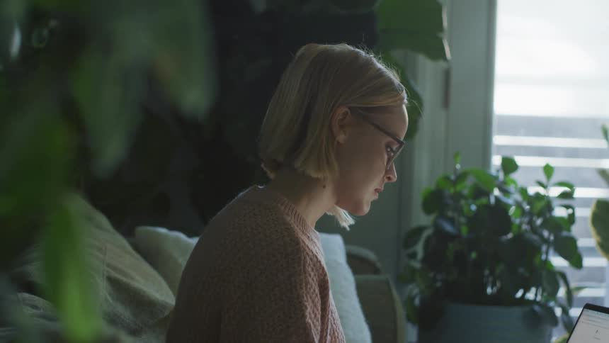 Woman wearing sweater sits on living room couch working on laptop