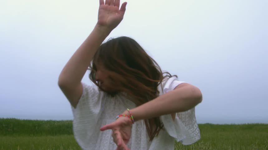 A woman in a white dress exuberantly dances and jumps around in an open field