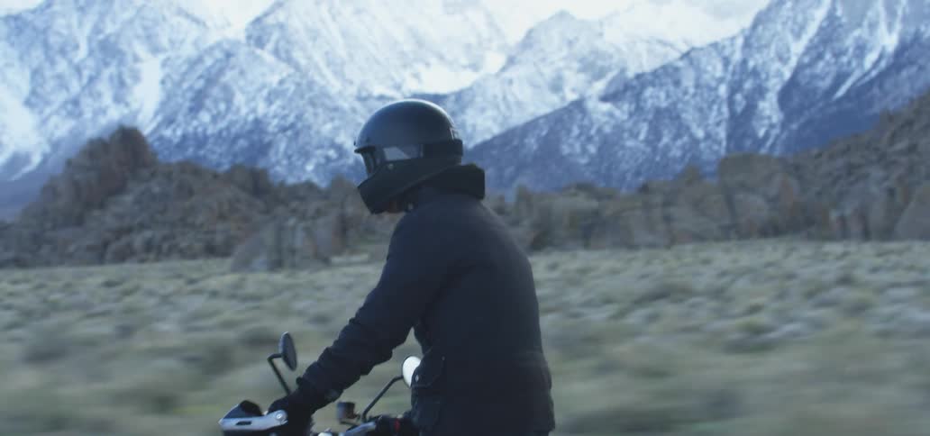 A man stands up while riding a motorcycle past a mountain range