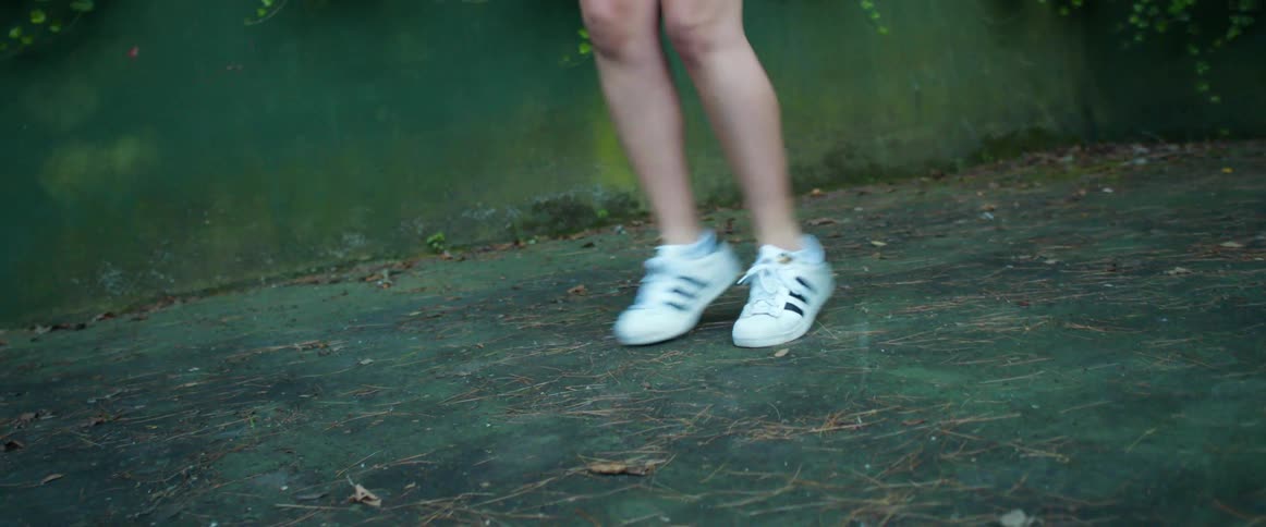 Woman wearing sneakers dances on concrete next to green wall partially covered in climbing vines