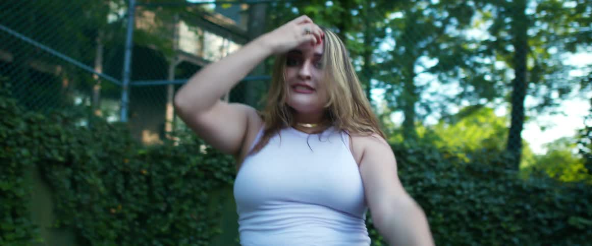 Blonde woman in a sleeveless top dances outdoors next to a fence and trees