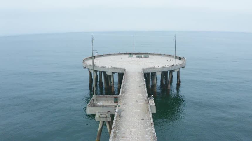A round observation deck with benches sits at the end of a long pier in the ocean