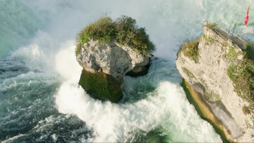 A rocky platform with stairs banister and guard railing with a swiss flag on a river next to a waterfall