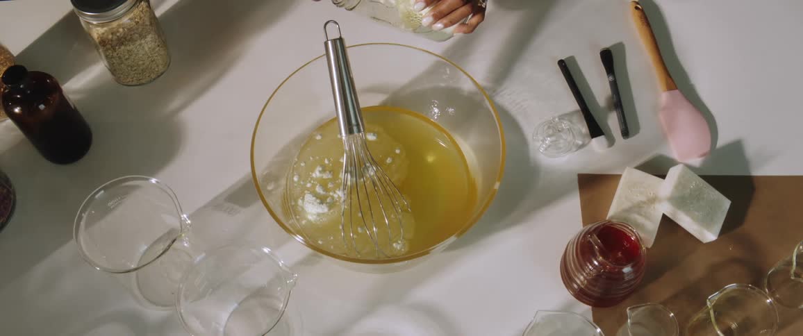A woman scoops a powder with a tablespoon into a liquid mixture in a bowl with a whisk on a kitchen counter