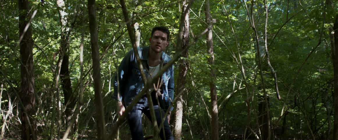 Bearded man in a denim jacket crouches as he walks through the forest