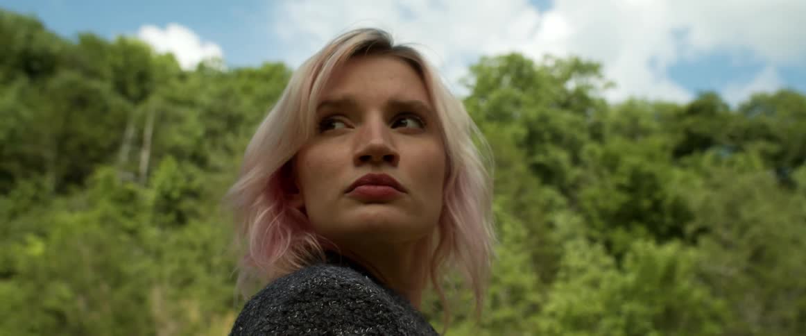 A woman standing in woodland during the day turns to look over her shoulder before turning back