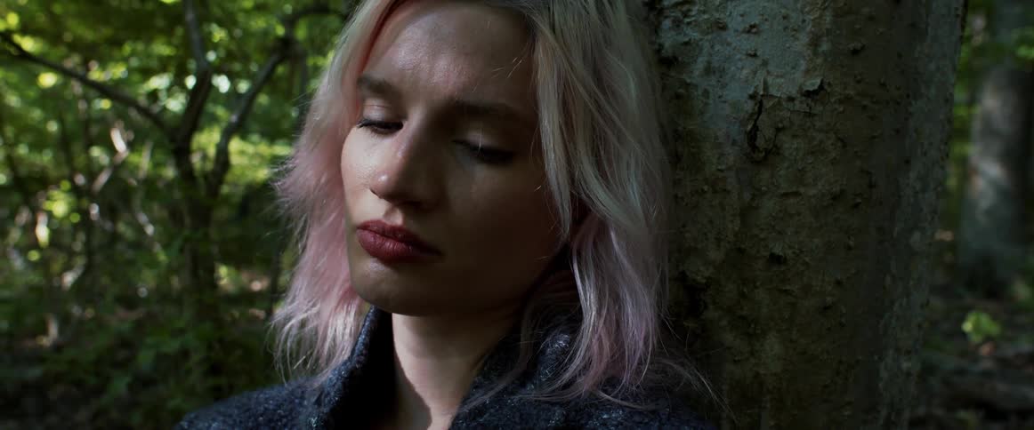 A depressed woman sits against a tree in a woodland during the day